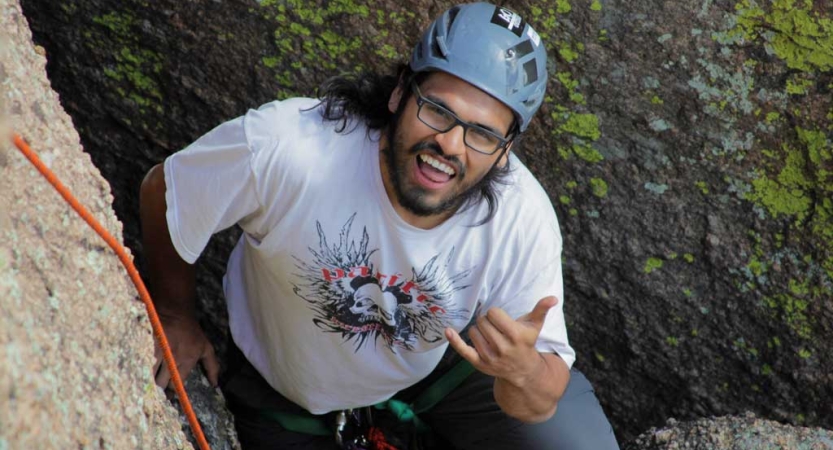 From above, a person wearing safety gear pauses to smile and give a "hang loose" sign as they climb towards the camera. 